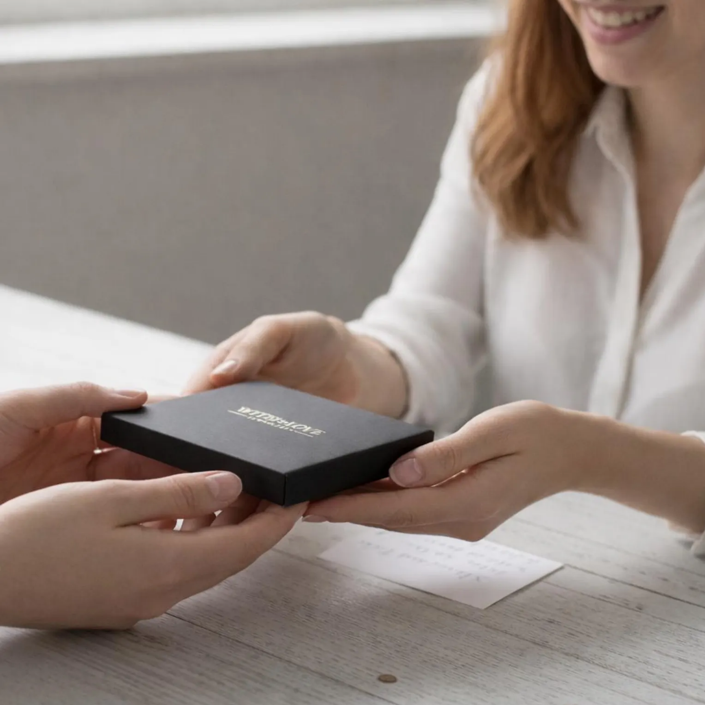 Person receiving a black box with 'With Love Jewellery UK' branding from another person on a light wooden surface.
