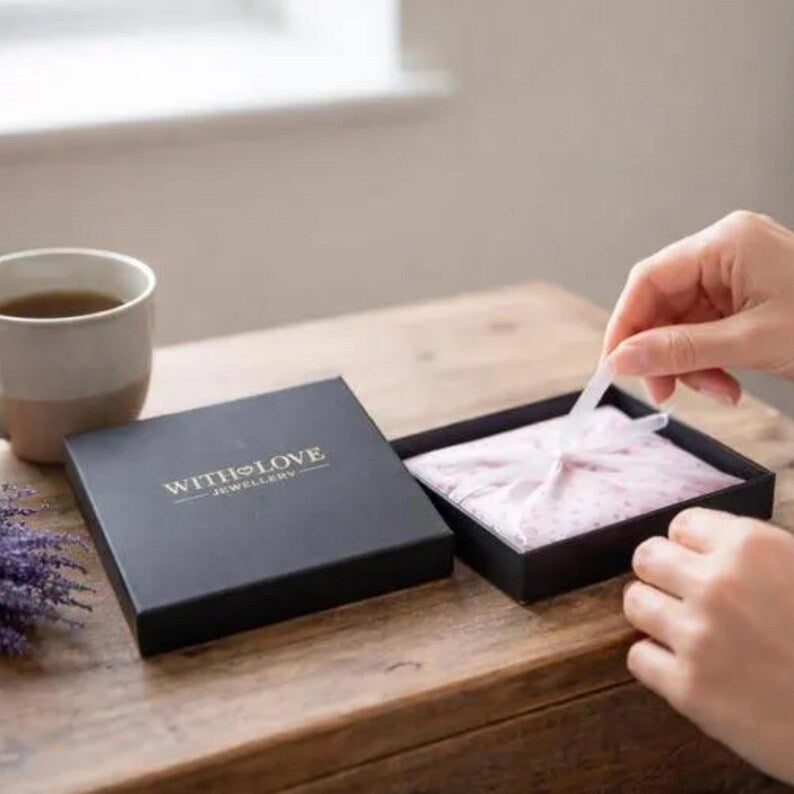 Person handling jewelry in a 'WITH LOVE' branded box on a wooden surface with a cup of tea and lavender.