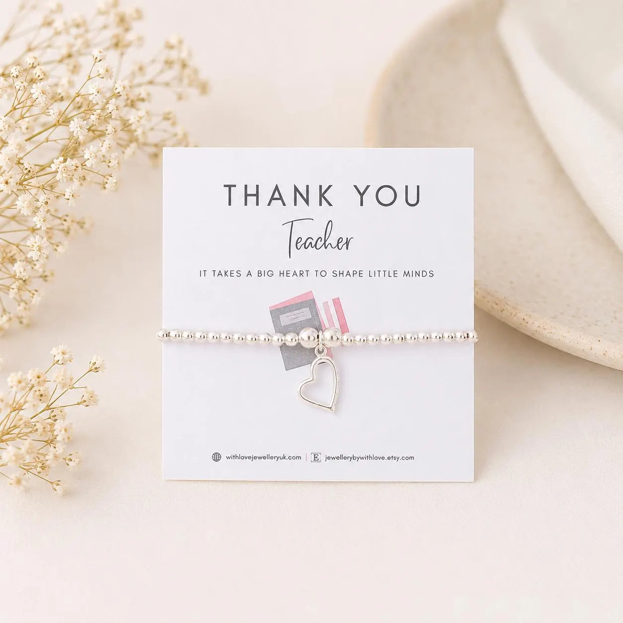 Bracelet with heart charm on a 'Thank You Teacher' card with flowers and plate in the background