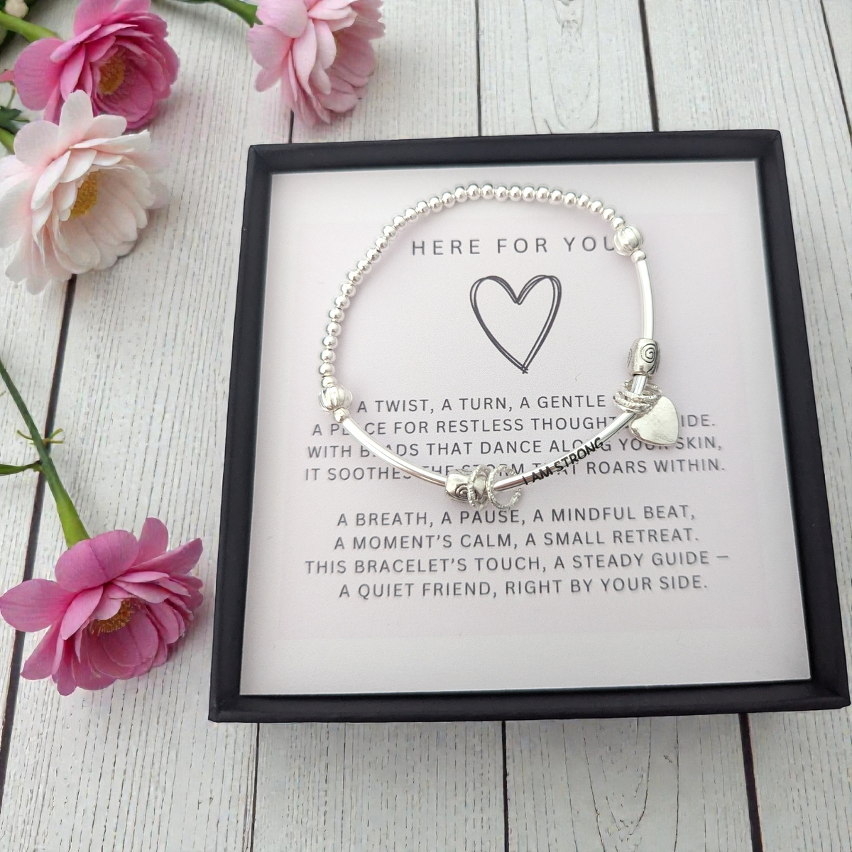 Silver bracelet with heart charm on a card with text, surrounded by pink flowers on a wooden surface