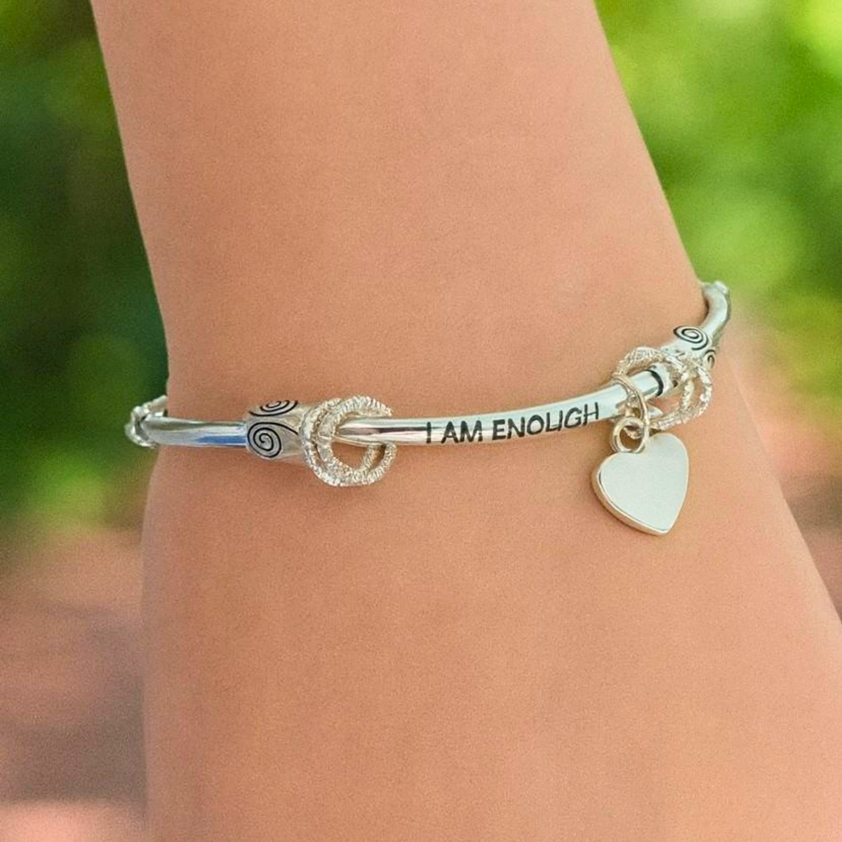 Silver bracelet with 'I AM ENOUGH' charm on a wrist against a blurred green background