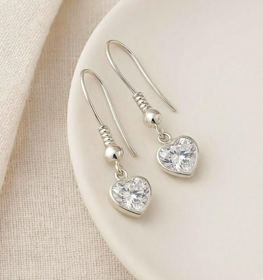 Silver heart-shaped earrings with clear gemstones on a beige surface