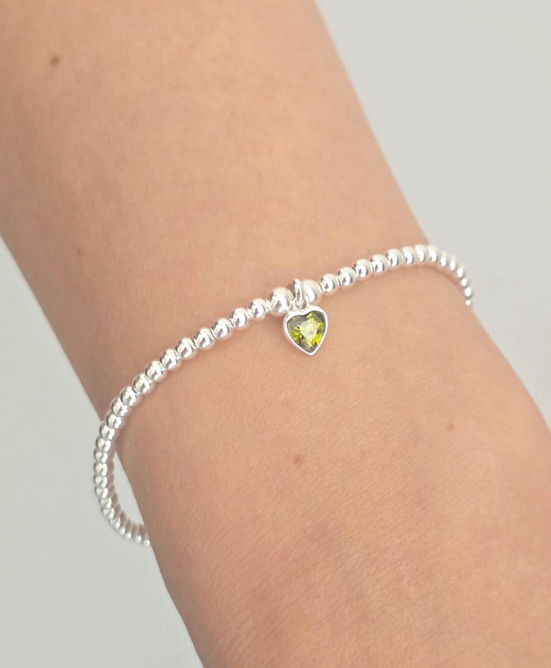 A delicate silver beaded bracelet with a small heart-shaped yellow gemstone charm, shown worn on a person's wrist against a light gray background.