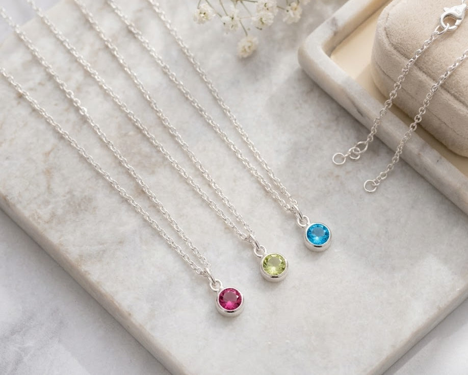 Silver necklaces with birthstone pendants on a marble surface with flowers.