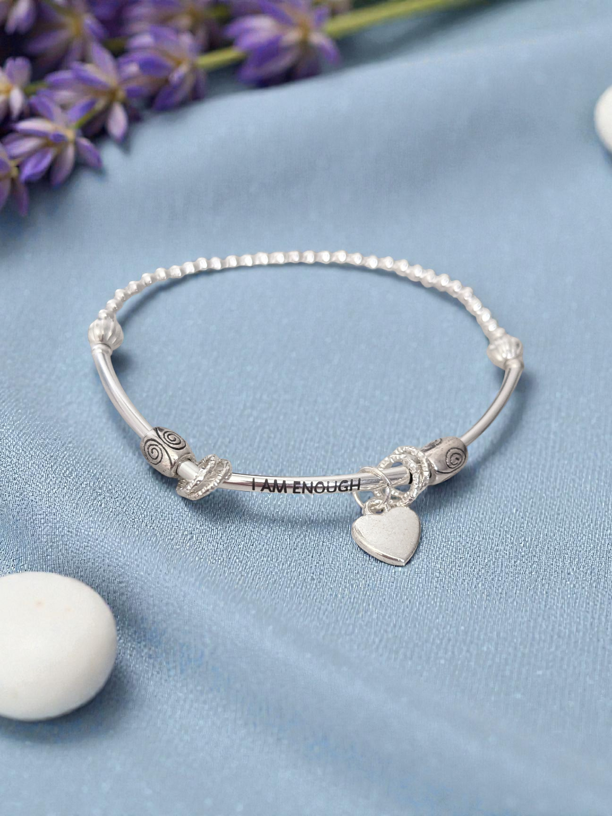 Silver bracelet with heart charm and 'I AM ENOUGH' message on a blue surface with lavender flowers.