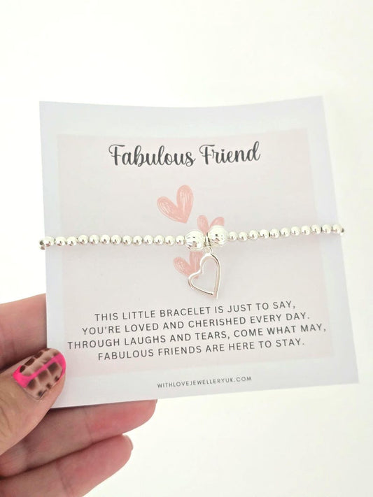 A silver beaded bracelet with a dangling heart charm displayed on a white gift card held by a hand. The card is printed with the words 'Fabulous Friend' and a sentimental message.