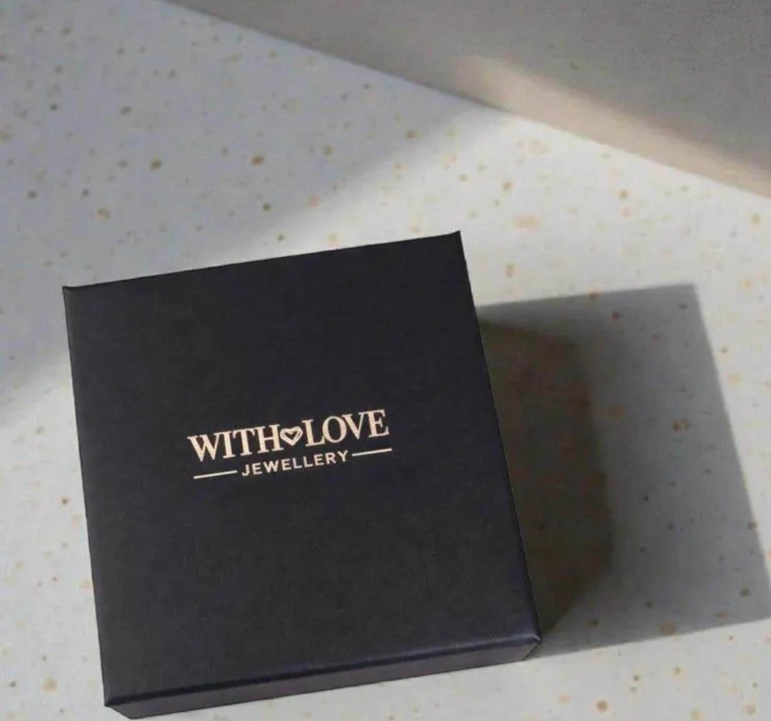 A black square jewelry box with gold foil branding reading 'WITH LOVE JEWELLERY' on a speckled light countertop.