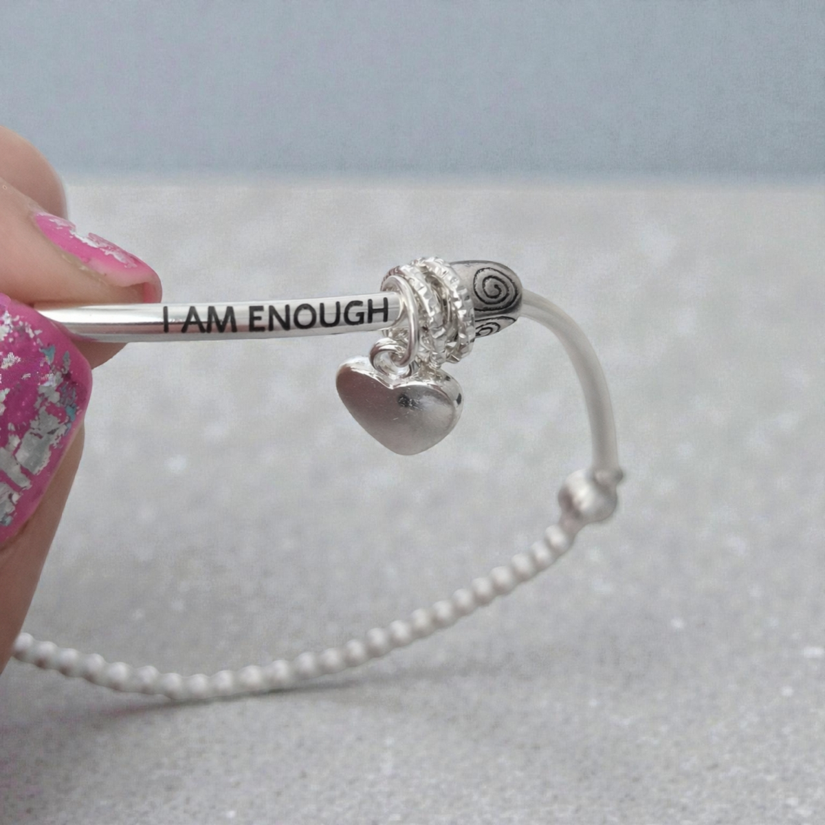 Silver bracelet with 'I AM ENOUGH' charm held by a hand on a light gray background