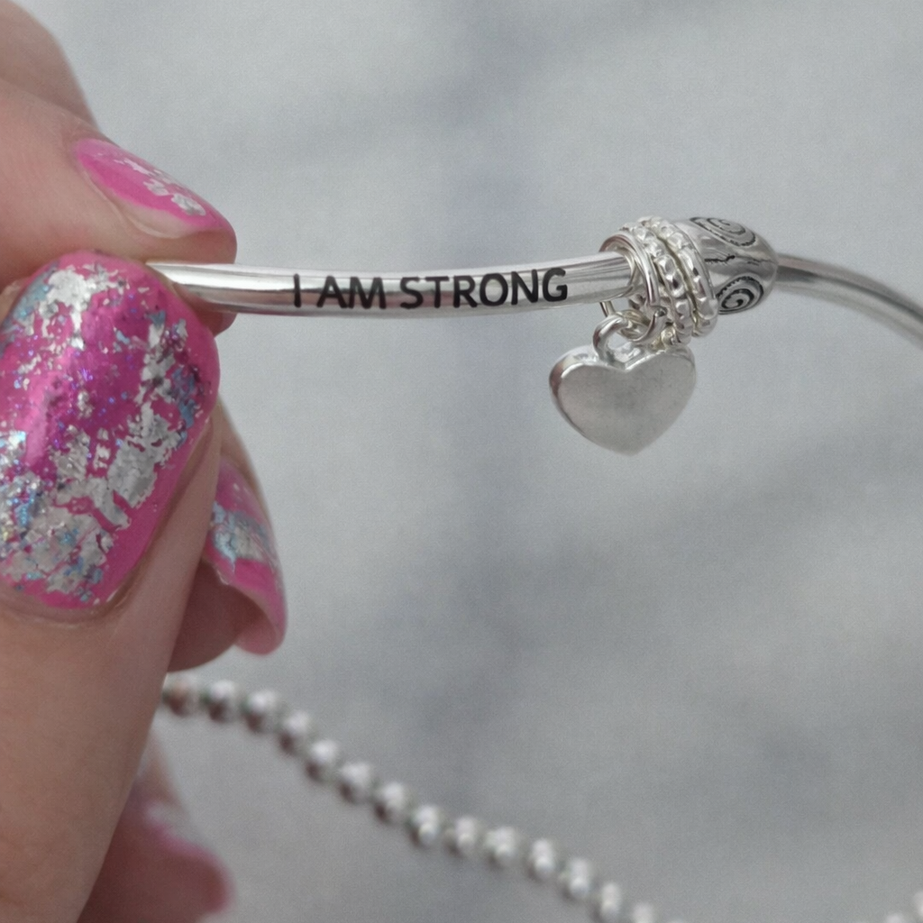 Silver bracelet with 'I AM STRONG' engraving held by a hand with pink nail polish.