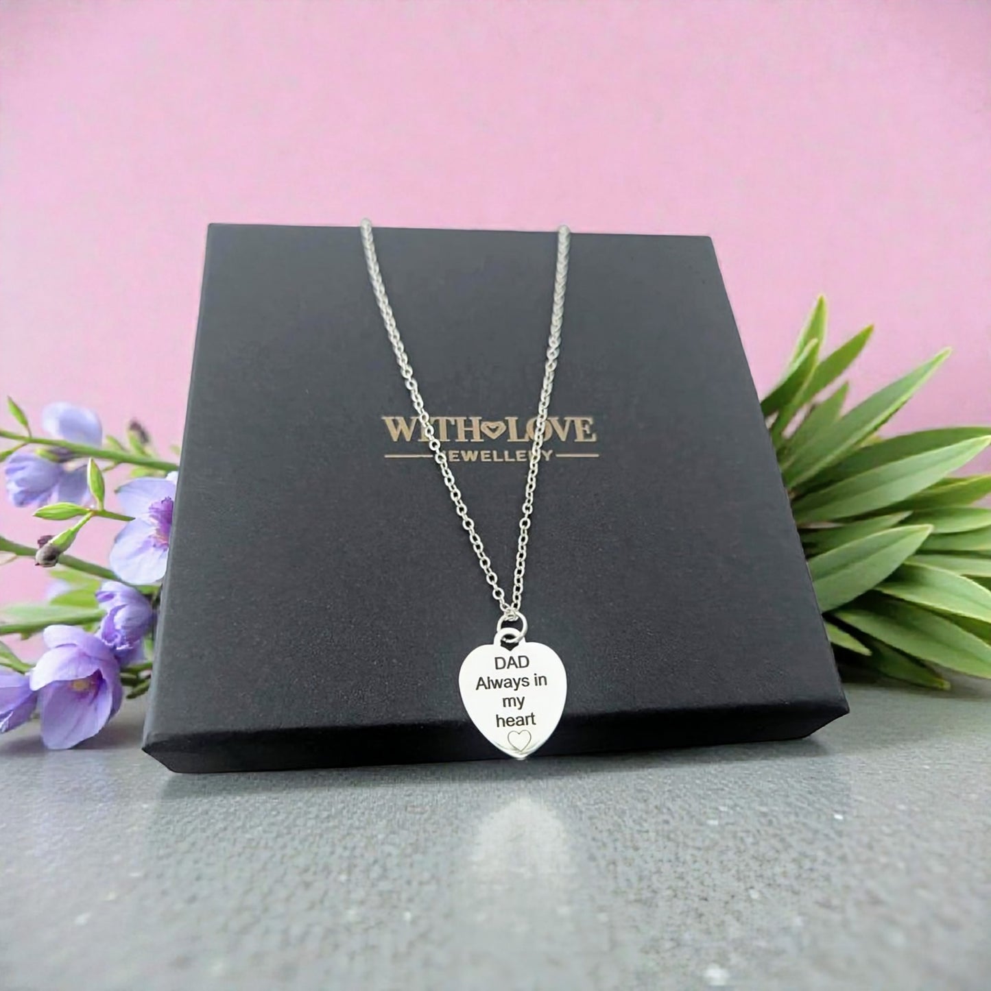 Silver necklace with a heart-shaped pendant on a black box with 'With Love Jewellery' branding, against a pink background with flowers and greenery.