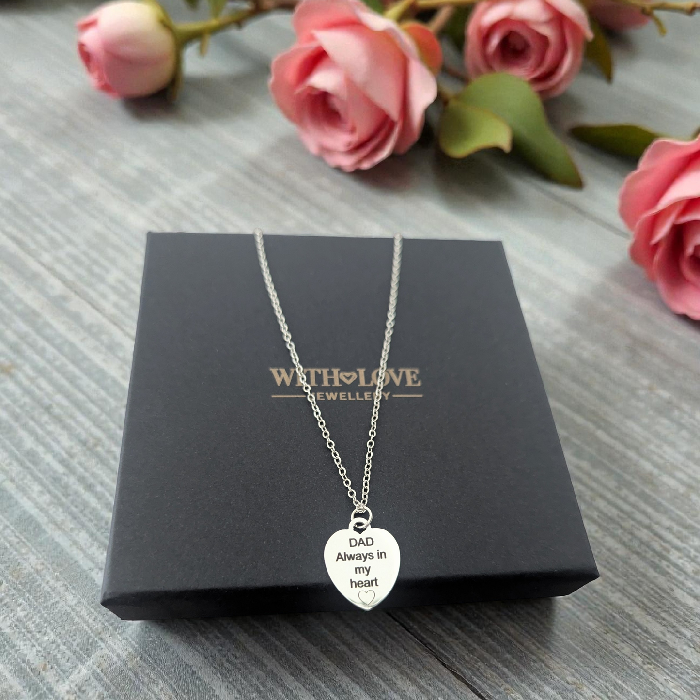 Silver necklace with a heart-shaped pendant on a 'With Love' jewelry box with pink flowers in the background.