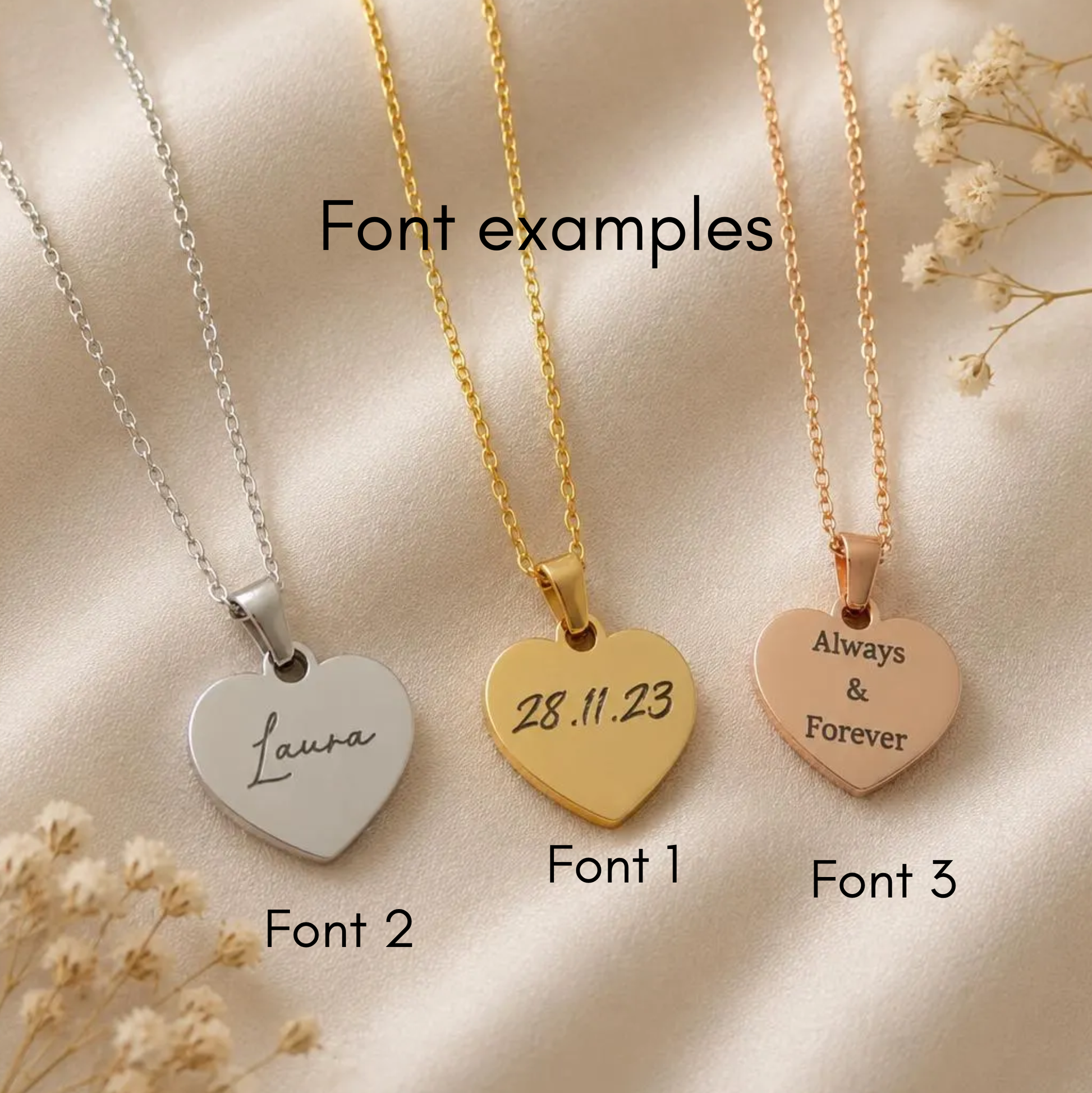 Three heart-shaped pendant necklaces displayed on a cream satin fabric with dried flowers. The left necklace is silver with the name 'Laura' engraved, labeled Font 2.