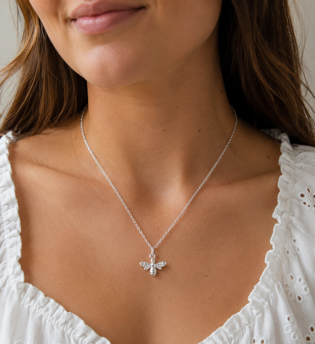 A close-up of a woman wearing a delicate silver chain necklace with a small bee pendant adorned with tiny clear crystals, paired with a white eyelet top.