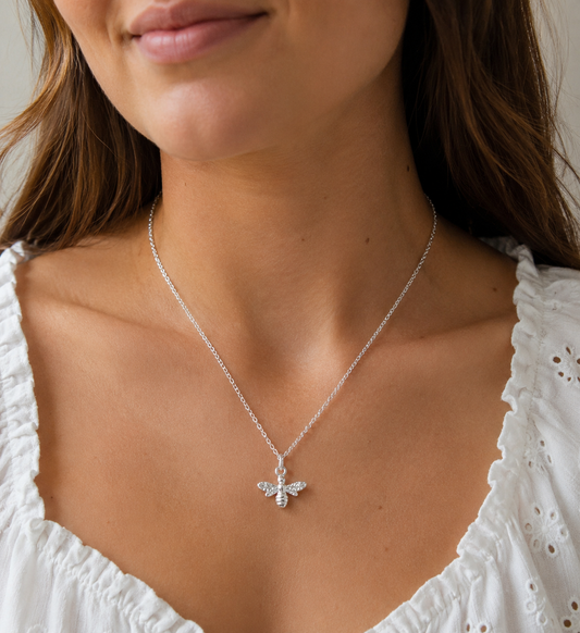 A close-up of a woman wearing a delicate silver chain necklace with a small bee pendant adorned with tiny clear crystals, paired with a white eyelet top.
