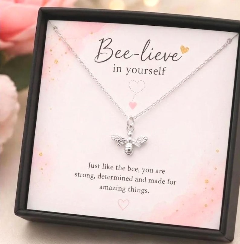 A silver bee pendant necklace displayed on a white card inside a black gift box. The card reads 'Bee-lieve in yourself' and features a soft pink watercolor design with gold speckles.