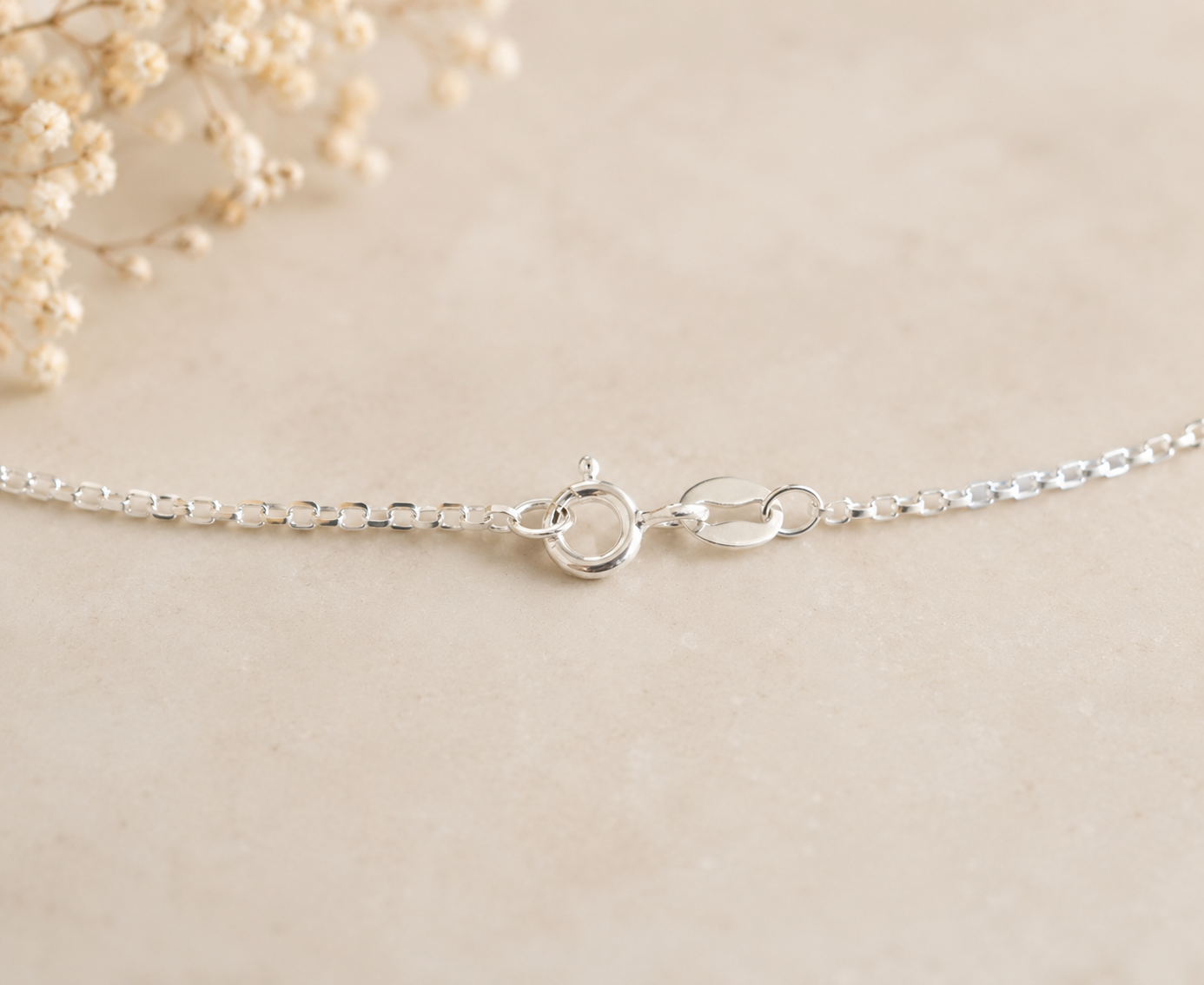 A close-up of a delicate silver chain bracelet showing its lobster clasp and oval links, laid flat on a light beige textured surface with dried baby's breath flowers in the top-left corner.