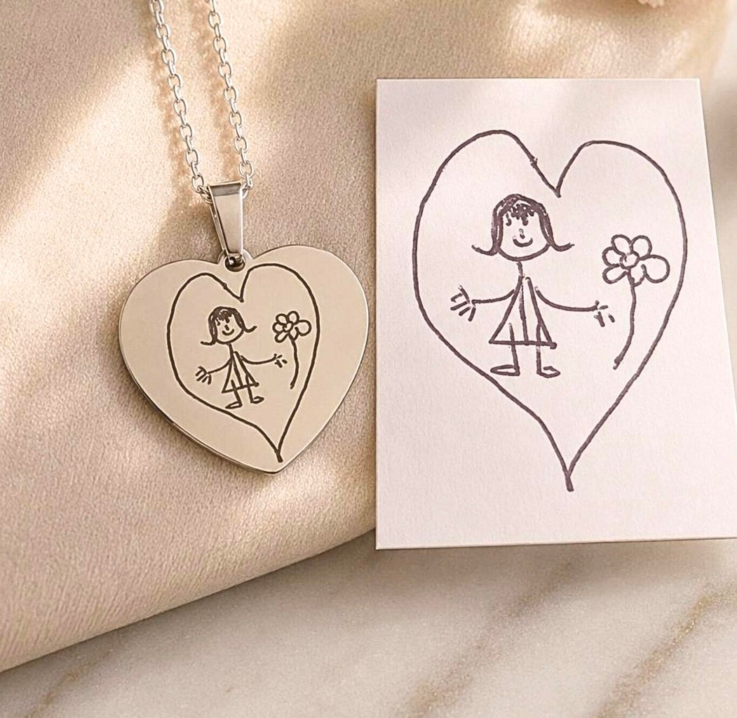 A heart-shaped pendant necklace with a silver-toned chain, featuring an engraved childlike figure and flower inside a heart, displayed on a soft beige fabric next to a white card with a hand-drawn matching heart illustration.