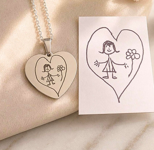 A heart-shaped pendant necklace with a silver-toned chain, featuring an engraved childlike figure and flower inside a heart, displayed on a soft beige fabric next to a white card with a hand-drawn matching heart illustration.