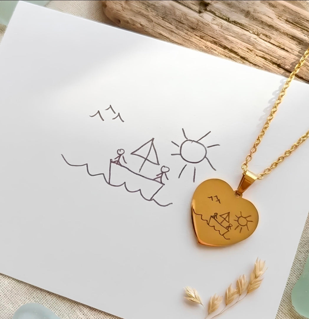 A gold heart-shaped pendant necklace with a hand-drawn beach scene, displayed on a white card next to a matching gold chain. The card features a simple sketch of a sailboat, sun, and birds, styled with dried wheat stalks and a piece of wood in the background.