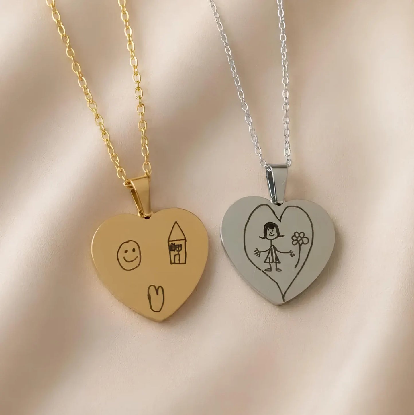 Two heart-shaped pendant necklaces displayed on a soft beige fabric background. One necklace is gold with a gold chain and a gold heart pendant engraved with simple doodles.