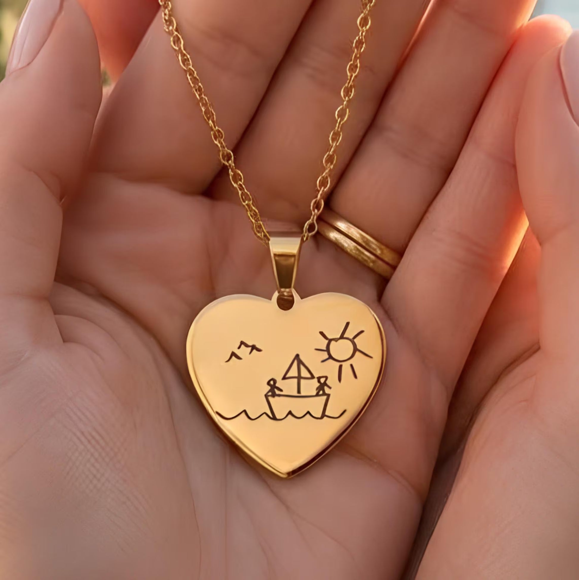 A gold heart-shaped pendant necklace with a simple boat, sun, and bird engraving, held in a person's hand.