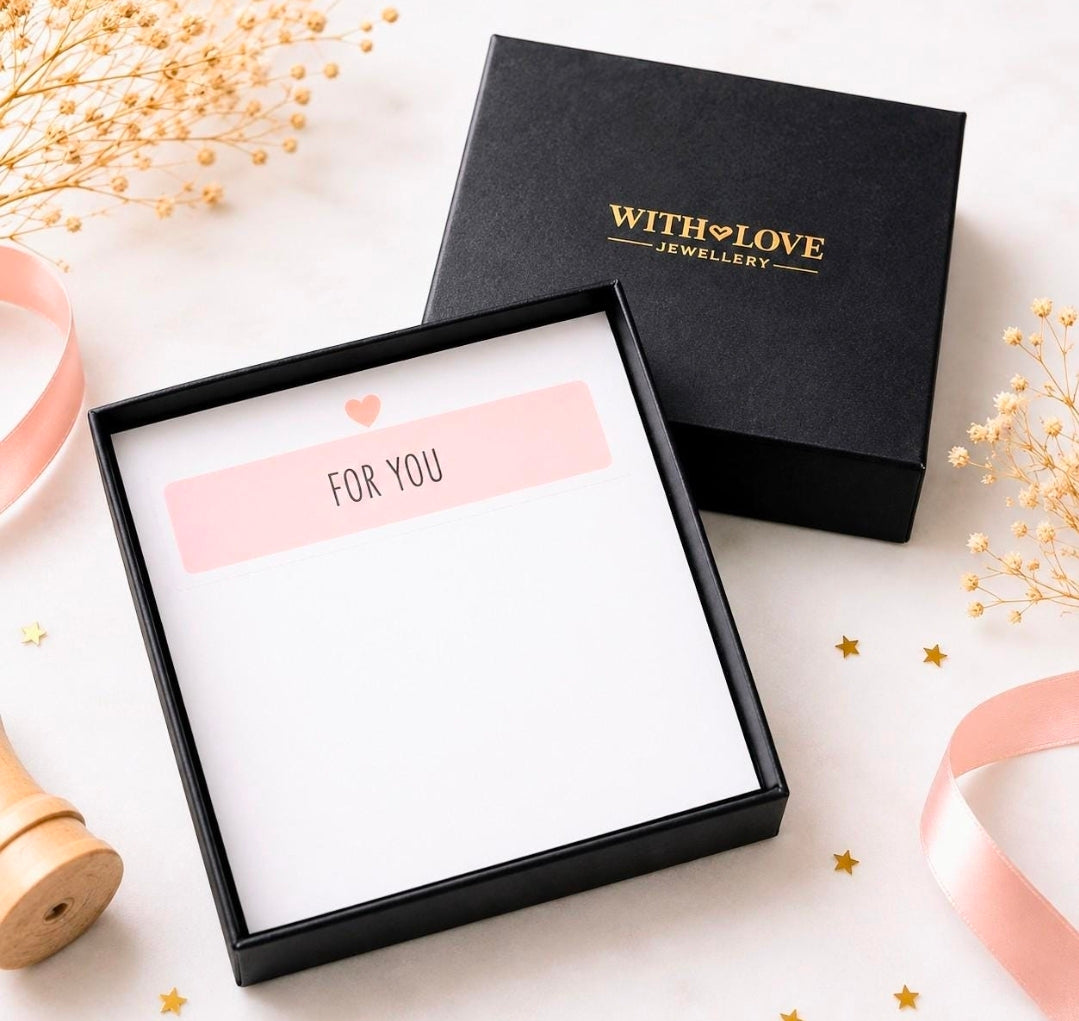 A black gift box set from WITH LOVE JEWELLERY, featuring a black outer box with gold lettering and an open inner box containing a white card with a pink 'FOR YOU' label. The scene is styled with pink ribbons, dried flowers, and gold star confetti on a light background.