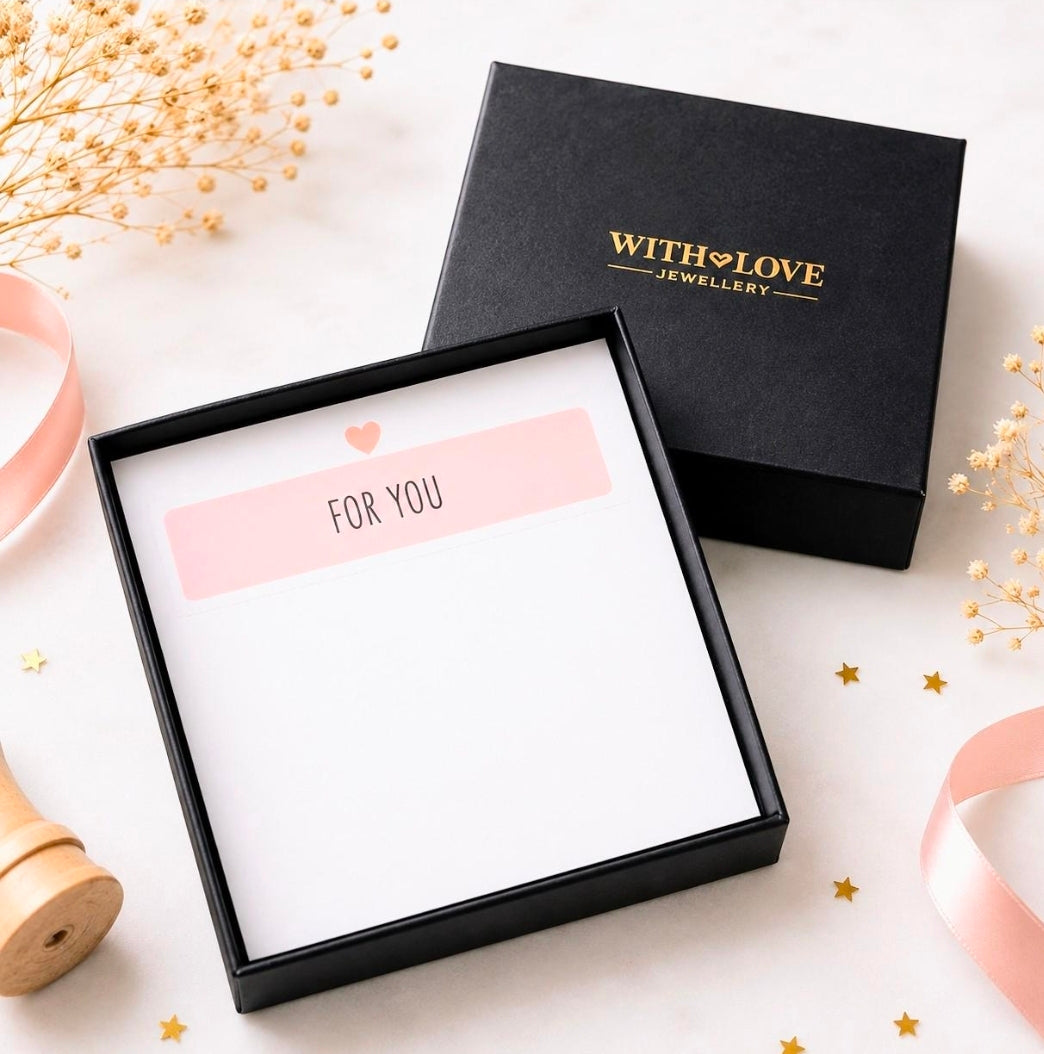 A black gift box set with a white insert card reading 'FOR YOU' and a small pink heart, shown next to the closed lid printed with 'WITH LOVE JEWELLERY' in gold. Styled on a light surface with dried flowers, a pink ribbon, and small gold stars.