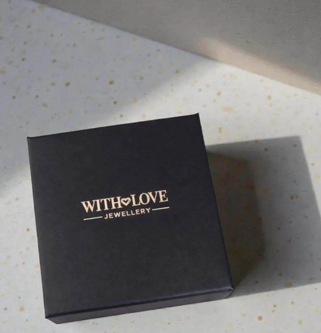 A square black jewelry box with gold foil branding that reads 'WITH LOVE JEWELLERY' on a speckled light beige countertop.