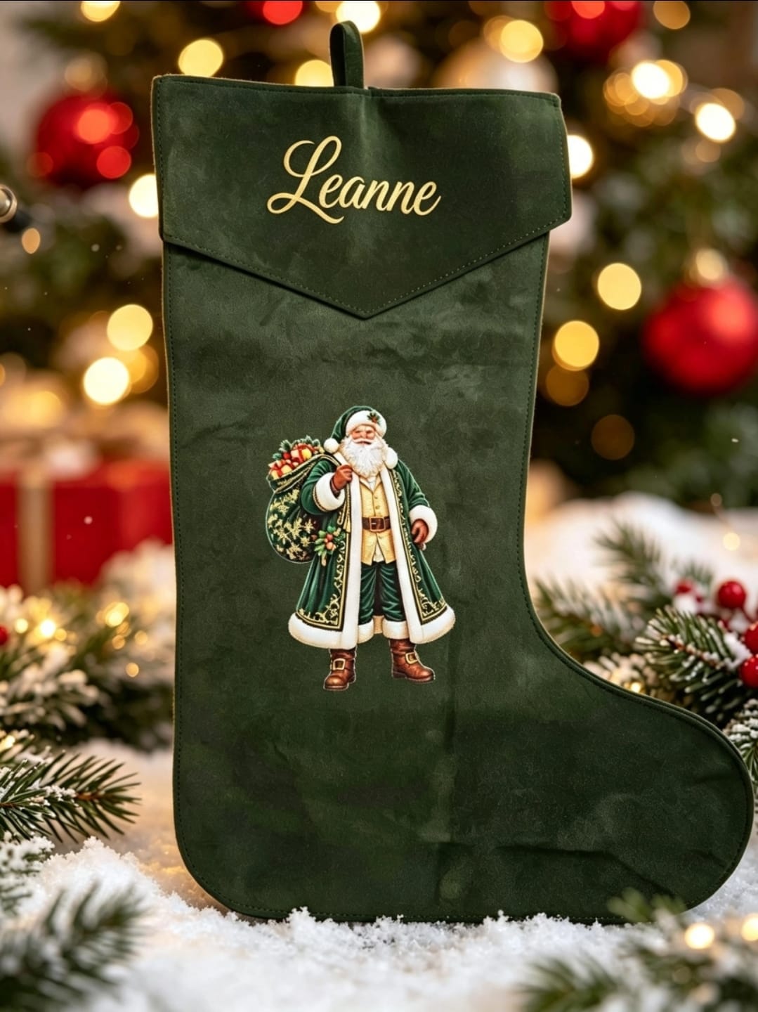 A dark green velvet Christmas stocking with gold cursive embroidery reading 'Leanne' and an embroidered Santa Claus figure, set against a festive blurred holiday background with lights, ornaments, pine branches, and snow.