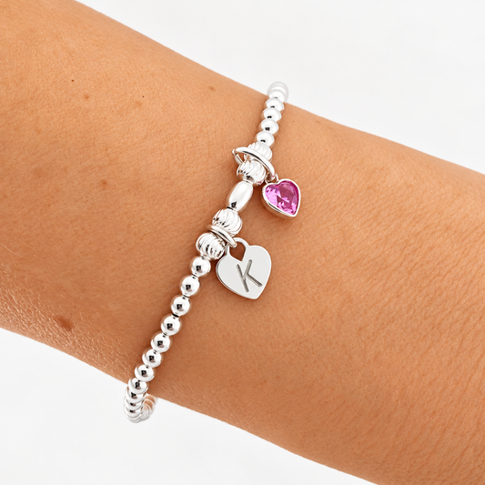 A silver beaded stretch bracelet with two heart charms, one engraved with the letter 'K' and the other featuring a pink heart-shaped stone, shown on a person's wrist.