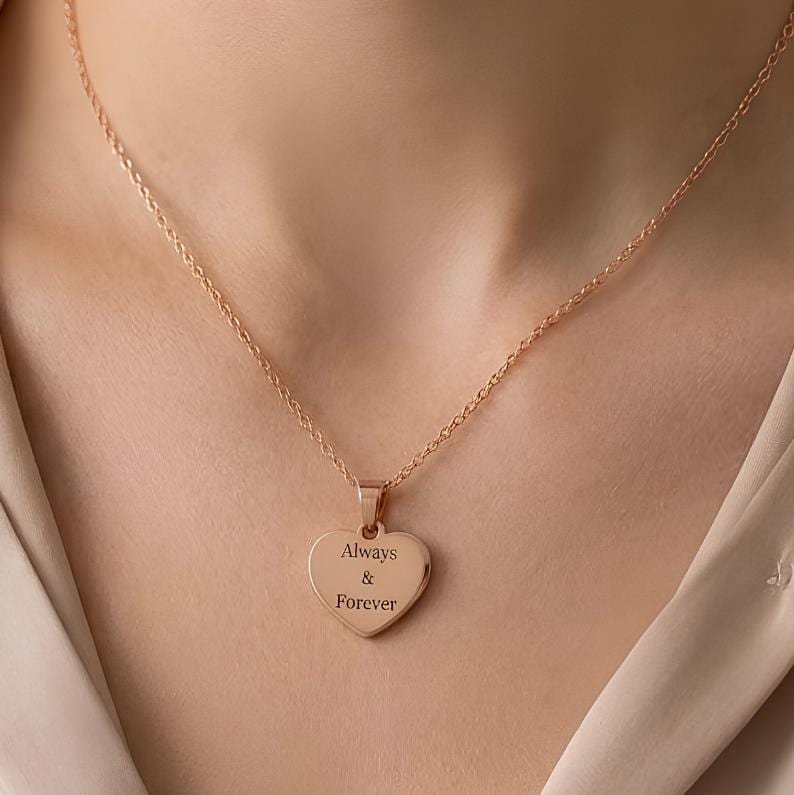 A close-up of a person's neck and chest wearing a delicate rose gold chain necklace with a heart-shaped pendant engraved with the words 'Always & Forever'.
