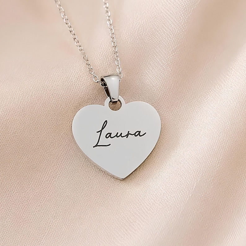 A silver heart-shaped pendant necklace engraved with the name 'Laura' in black script, resting on a soft, light pink satin fabric background.