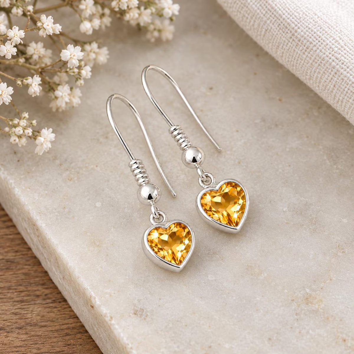 A pair of silver heart-shaped drop earrings with faceted yellow gemstones, displayed on a light beige stone surface with small white flowers and a textured fabric in the background.
