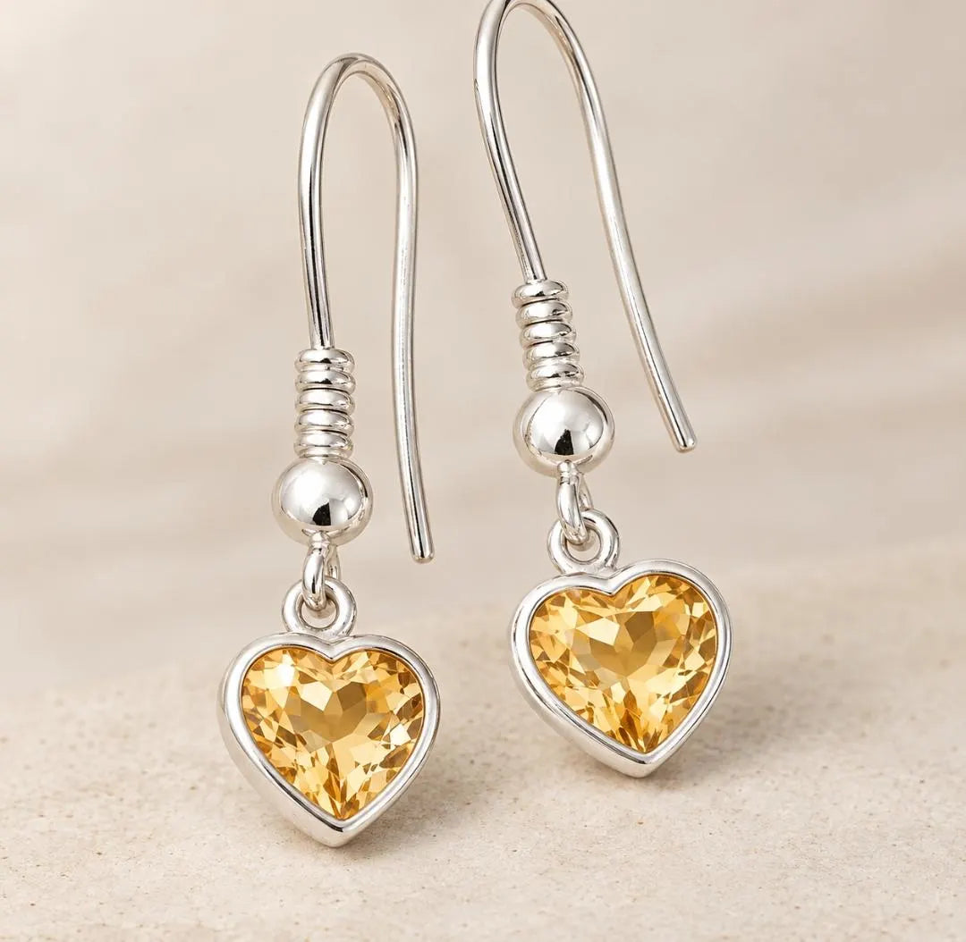 A pair of silver heart-shaped drop earrings with faceted yellow gemstones, displayed on a light beige textured background.
