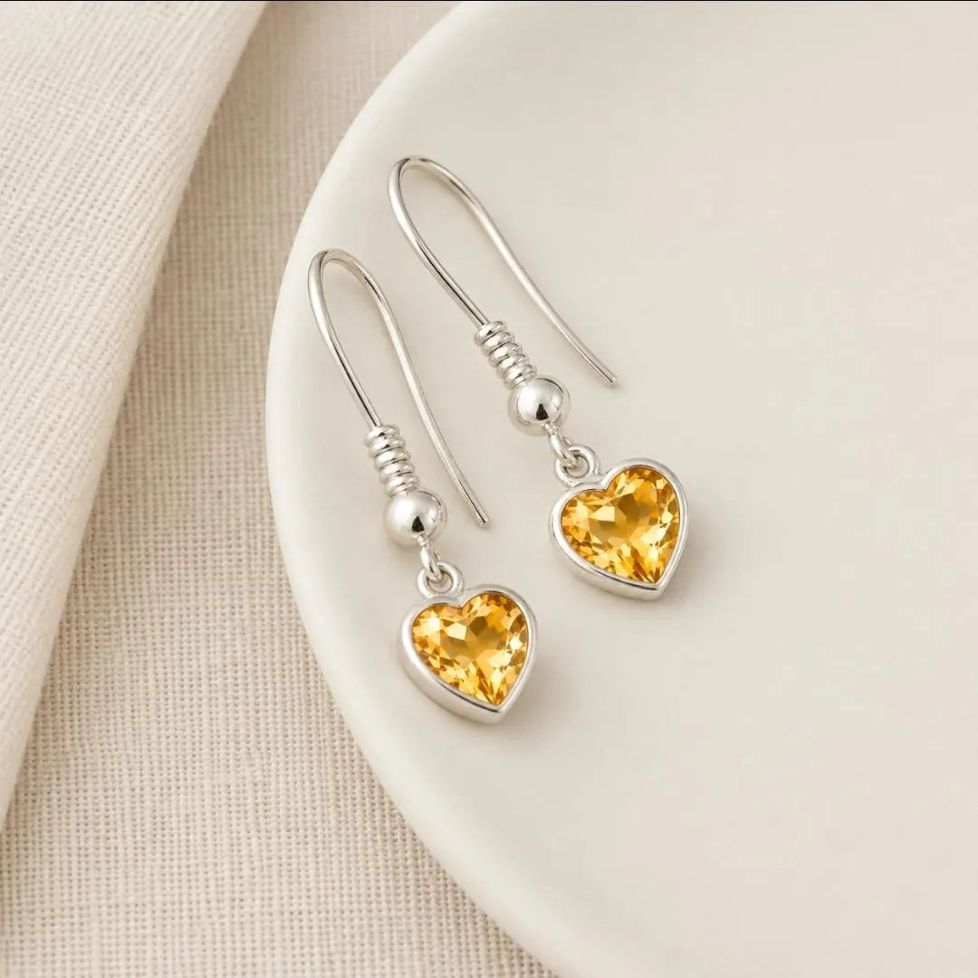 A pair of silver heart-shaped drop earrings with yellow gemstones, displayed on a light beige ceramic plate over a textured beige fabric background.