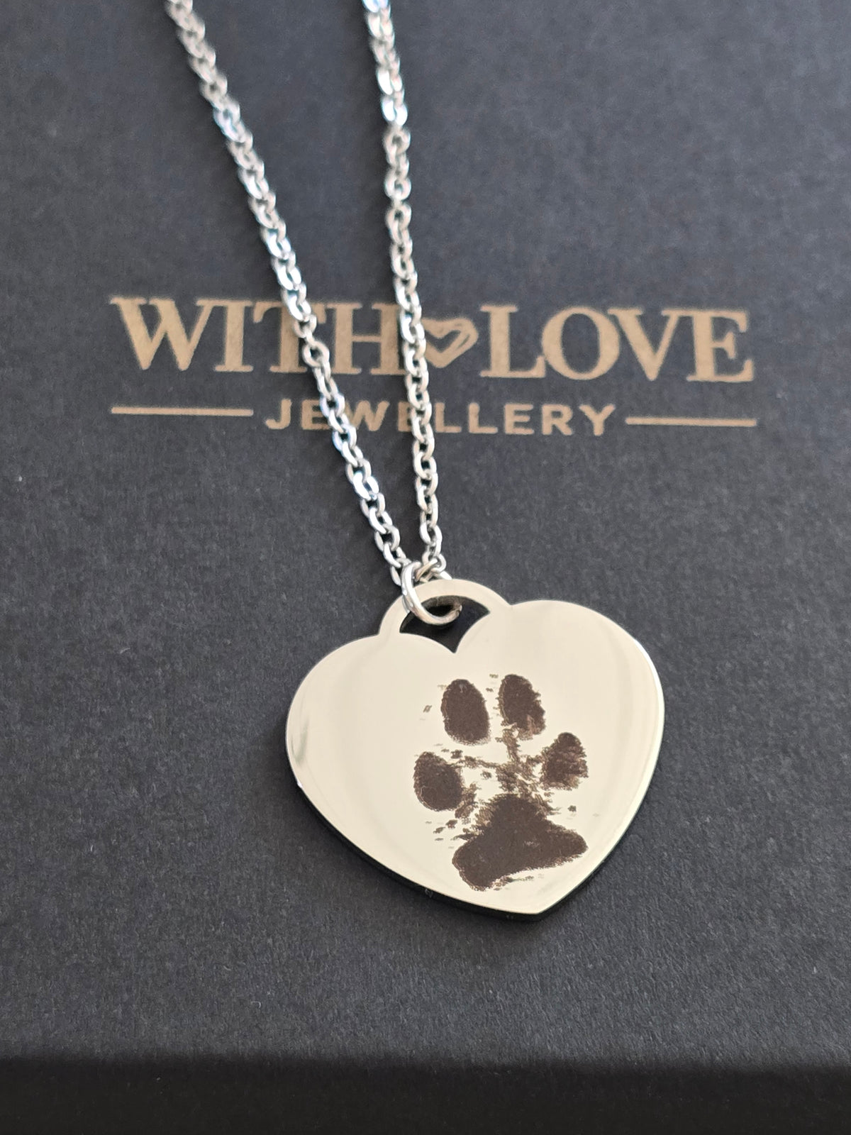 A silver heart-shaped pendant necklace with a dark brown paw print imprint, resting on a dark gray box printed with 'WITH LOVE JEWELLERY'.