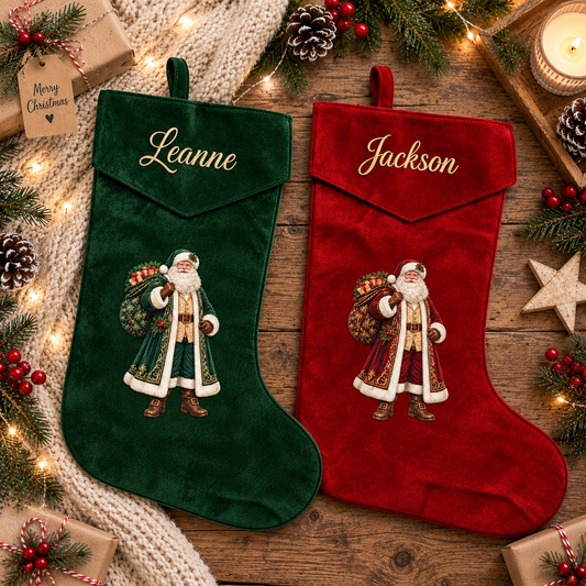 Two personalized Christmas stockings, one dark green velvet with the name 'Leanne' and one red velvet with the name 'Jackson', both featuring embroidered Santa Claus designs. They are displayed on a rustic wooden table surrounded by holiday decorations including pine branches, a knitted blanket, wrapped gifts, and a lit candle.