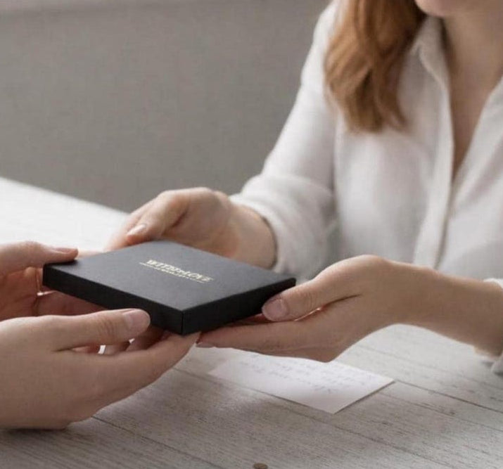 Person receiving a black box with a brand logo from another person on a light wooden surface.