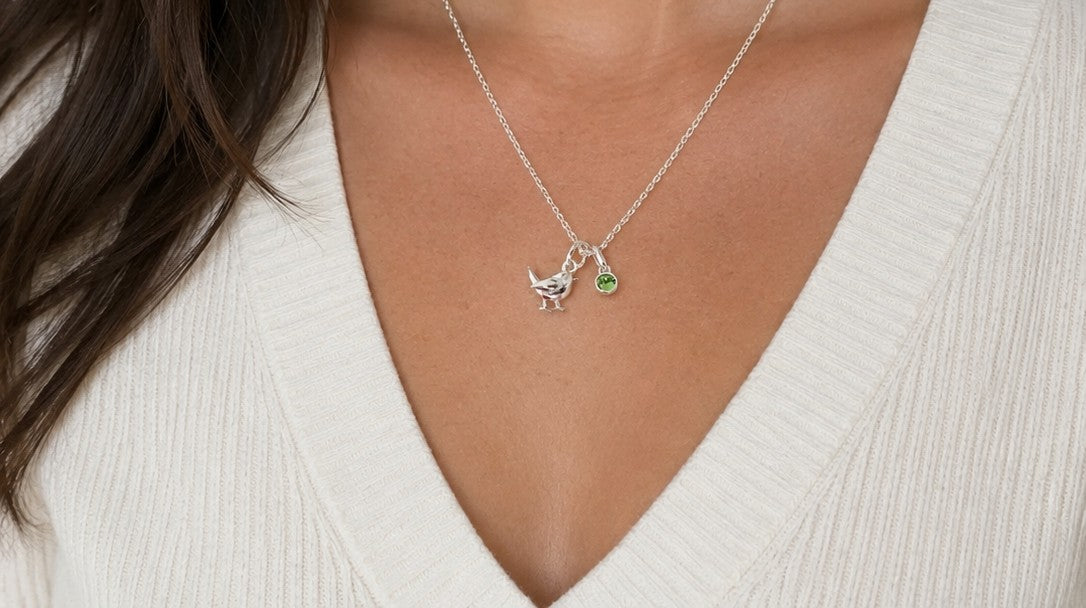 A close-up of a model wearing a delicate silver necklace with a small silver bird charm and a tiny green gemstone pendant, styled with a white ribbed V-neck top.