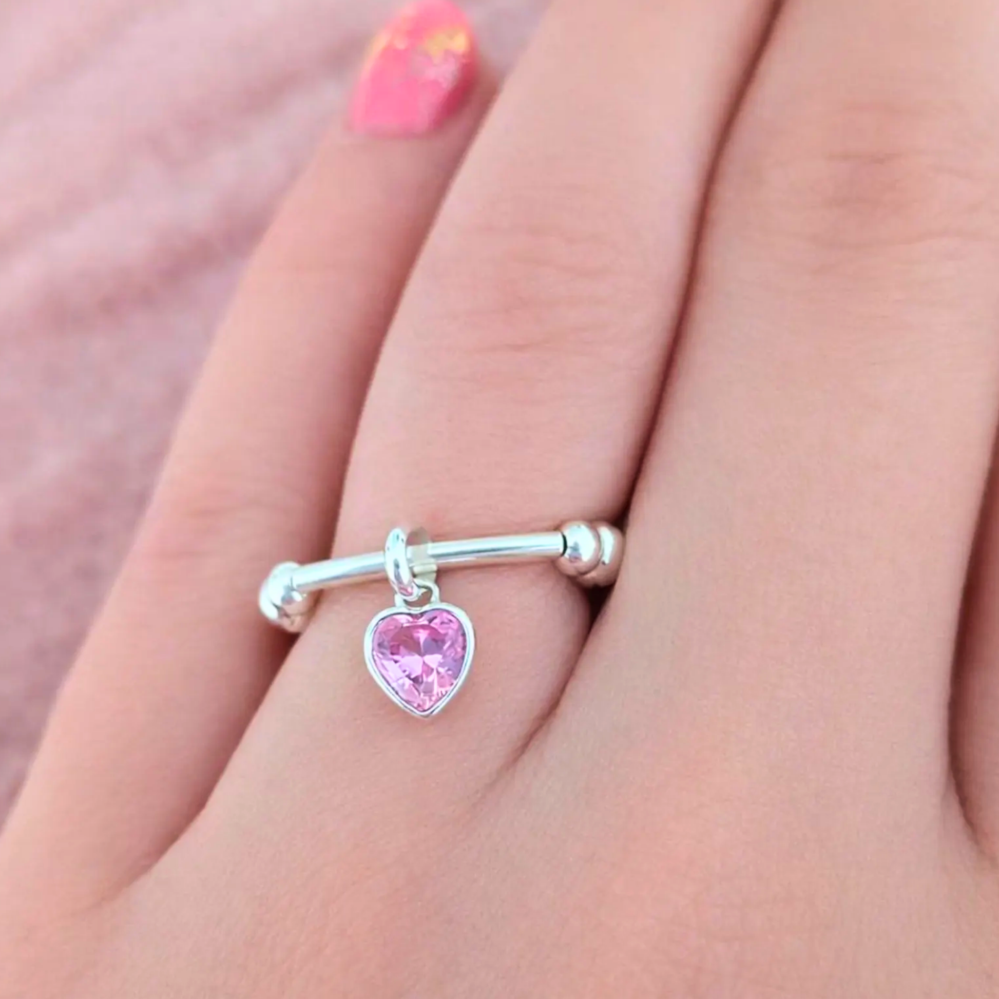 Silver ring with a pink heart charm on a person's finger against a pink background