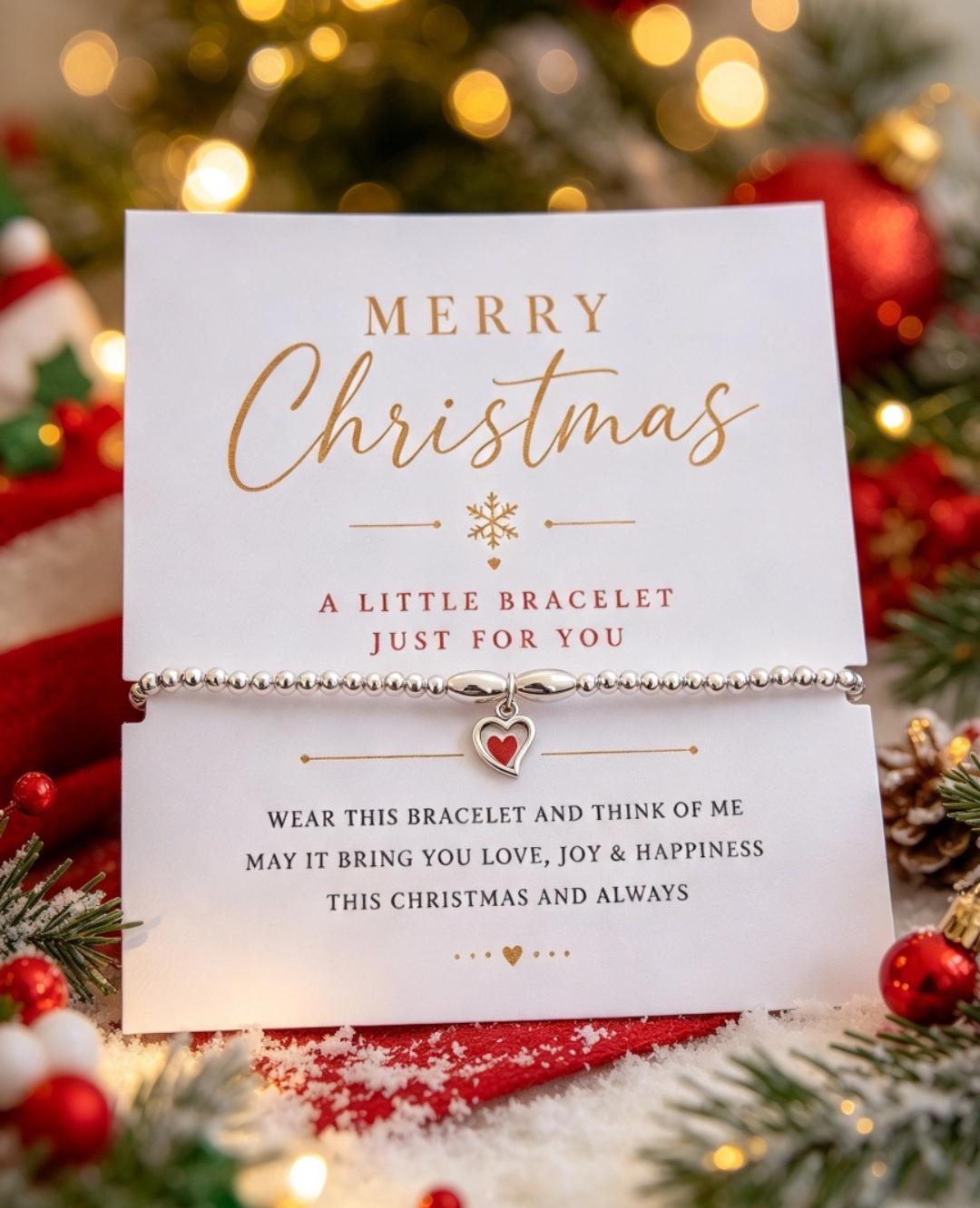 A silver bracelet with a small heart-shaped charm is displayed on a white gift card that reads 'Merry Christmas' and includes a sentimental message. The card is photographed in a festive holiday setting with red ornaments, green pine branches, and warm golden bokeh lights in the background.