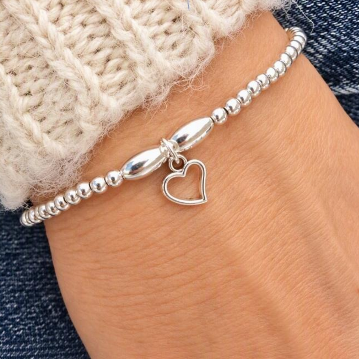 A silver beaded bracelet with a small open heart charm, shown worn on a wrist with a cream knit cuff and blue denim visible in the background.